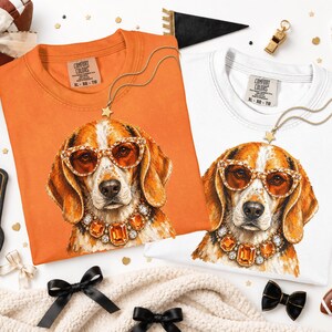 May include: Two Comfort Colors t-shirts, one orange and one white, each featuring a graphic print of a beagle dog wearing amber-colored sunglasses and a matching jeweled necklace. The shirts have a crew neckline and a tag printed on the inner collar.