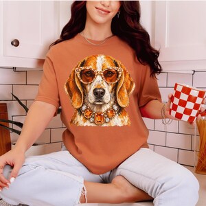 May include: A rust-colored short-sleeve t-shirt featuring a graphic print of a beagle dog wearing amber-colored sunglasses and a large, ornate necklace with orange and clear gemstones. The shirt is paired with light-wash denim jeans.