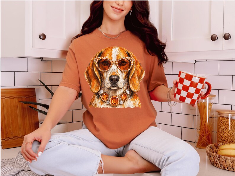 May include: A rust-colored short-sleeve t-shirt featuring a graphic print of a beagle dog wearing amber-colored sunglasses and a large, ornate necklace with orange and clear gemstones. The shirt is paired with light-wash denim jeans.