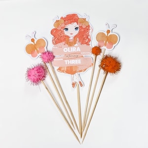 May include: Cake toppers for a birthday celebration. The set includes a girl illustration, butterflies, and pom-poms in shades of orange and pink. The text on the banner reads "OLIRA is turning THREE."
