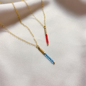 May include: Two delicate gold necklaces with pendant beads. One necklace features a strand of red beads, and the other has blue beads. The necklaces are displayed on a white satin background.