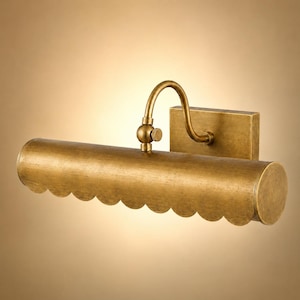 Op de afbeelding: Een wandlamp voor schilderijen met een goudkleurige metalen afwerking. Het armatuur heeft een horizontale cilindrische kap met een geschulpte onderrand, verbonden met een rechthoekige muurplaat via een gebogen arm met scharnier.
