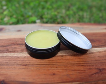 Può includere: Un piccolo contenitore di latta con un coperchio, riempito di un balsamo o unguento giallo. Il contenitore è su una superficie di legno.