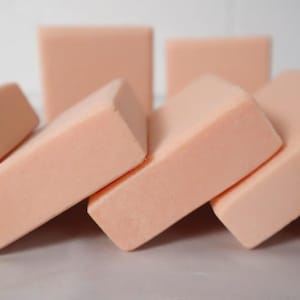 May include: Six rectangular bars of soap, all the same size and shape, are arranged in a row. The bars are a light peach color and have a smooth, matte finish.