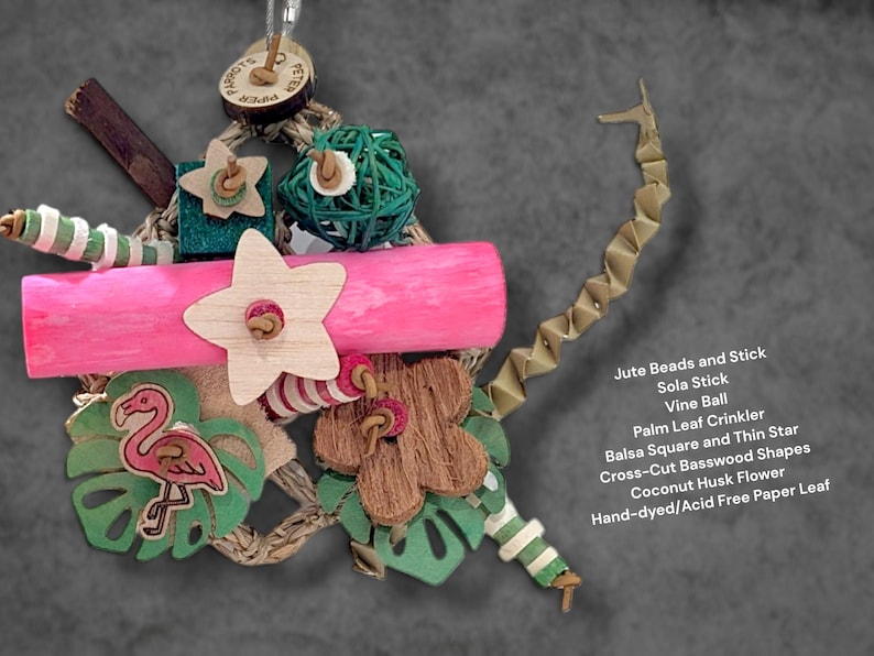 May include: A bird toy featuring a pink wooden cylinder, a green vine ball, a coconut husk flower, a wooden star, and a flamingo cutout. It includes jute beads, a sola stick, a palm leaf crinkler, balsa squares, and basswood shapes on a jute rope.