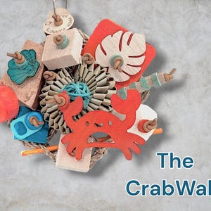 May include: A bird toy titled The CrabWalk featuring a central red wooden crab cutout, a pleated paper flower, a monstera leaf shape, and various colourful wooden blocks, beads, and teal shell accents strung together on a natural fibre rope base.