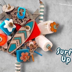 May include: A multi-textured bird toy featuring a central wooden surfboard shape with the text Surf's Up, surrounded by colourful wooden blocks, star-shaped pieces, and cylindrical beads strung on leather cords, with additional text Toys sold individually.