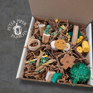 May include: A cardboard box filled with various parrot toys. The toys include wooden blocks, woven rings, and colorful, textured items. The box is lined with brown paper and features the "Peter Piper Parrots" logo in the upper left corner.