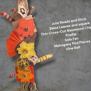May include: A bird toy featuring orange and yellow felt leaves, a bright orange sola fan, a yellow vine ball, mahogany pod pieces, balsa wood squares, and basswood chippers. Text reads: Jute Beads and Stick, Balsa Leaves and square, Thin Cross-Cut Basswood Chippers, Pradip, Sola Fan, Mahogany Pod Pieces, Vine Ball.