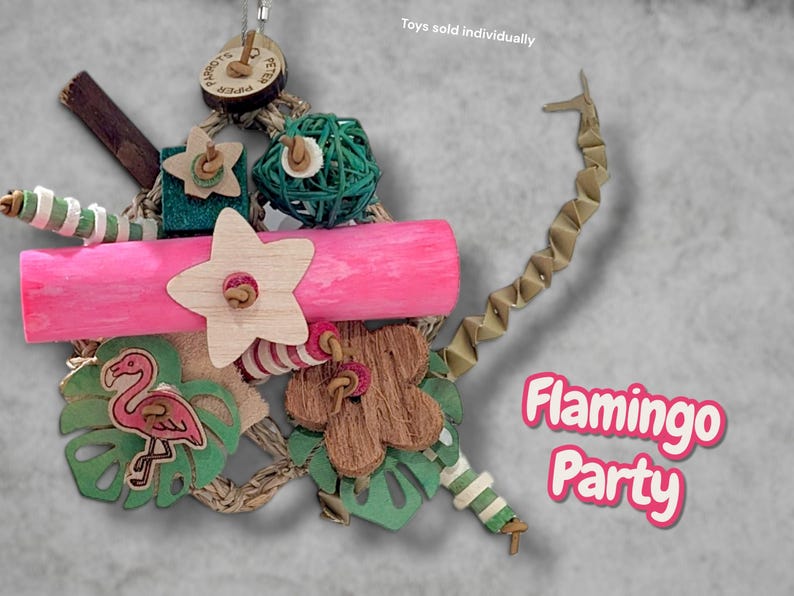 May include: A bird toy featuring a bright pink wooden log, a light-coloured wooden star, a green woven ball, and a flamingo cutout on a green leaf. It includes a brown flower shape, striped beads, and a zigzag paper strip. Text reads Flamingo Party.