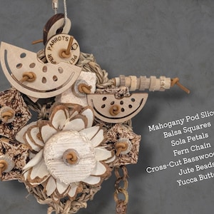 May include: A bird toy featuring mahogany pod slices, balsa squares, sola petals, a fern chain, cross-cut basswood shapes, jute beads, and a yucca button. The text lists these materials: Mahogany Pod Slices, Balsa Squares, Sola Petals, Fern Chain, Cross-Cut Basswood Shapes, Jute Beads, Yucca Button.