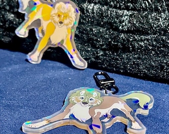 May include: Two iridescent keychains featuring cartoon wolf-like characters. The first keychain features a yellow wolf with a black nose and black eyes. The second keychain features a brown and white wolf with green fur around its neck and blue eyes.