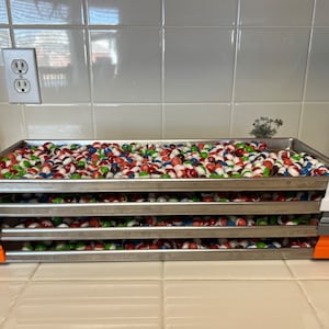 May include: Four silver baking sheets stacked on top of each other, filled with colorful candy. The sheets have orange, gray, and white clips on the sides.