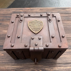 May include: A brown, rectangular chest with metal accents and a hinged lid. The chest features a brass emblem that reads "PROPERTY OF WELLS FARGO & CO EXPRESS SAN FRANCISCO DIVISION". The chest is secured with a metal latch and rivets.