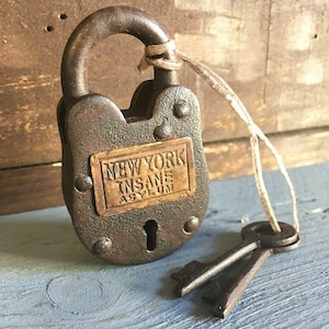 May include: A vintage, rusty padlock with the words "NEW YORK INSANE ASYLUM" on a brass plate. Two matching keys are attached with a string. The lock is a dark brown colour and appears aged.