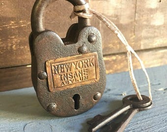 Puede incluir: Un candado oxidado y vintage con las palabras "NEW YORK INSANE ASYLUM" en una placa de latón. Dos llaves a juego están unidas con una cuerda. El candado es de color marrón oscuro y parece envejecido.