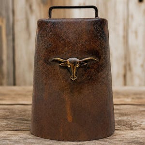 May include: A rustic, brown metal bell with a handle and a decorative brass bull head emblem. The bell has a weathered appearance, suggesting it is an antique or vintage item. The bell is likely used for decorative purposes or as a collectible item.
