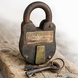 Puede incluir: Un candado antiguo y oxidado con una placa de latón que dice "FOLSOM STATE PRISON Folsom, California 1880". Dos llaves descansan debajo del candado sobre una superficie de madera. El candado tiene un asa grande y arqueada.