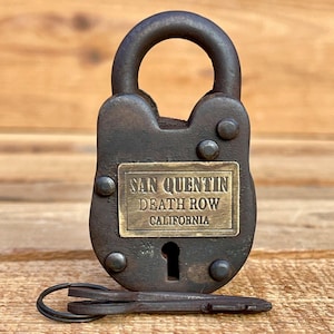 Puede incluir: Un candado vintage de metal oscuro con una llave. El candado tiene forma rectangular con un asa curva y una placa de latón que dice "SAN QUENTIN DEATH ROW CALIFORNIA". La llave está debajo.