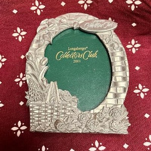 May include: A silver-colored metal picture frame with a green oval center. The frame has a floral and basket design with the words "Longaberger Collectors Club 2001" on the green background and a small tag.
