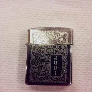 May include: Silver-toned metal lighter with intricate floral engraving. The lighter features the name "JODI" vertically engraved on the front. The design includes detailed scrollwork and a rectangular frame.