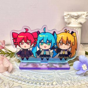 May include: A clear acrylic stand featuring three anime-style characters with red, blue, and yellow hair. The characters are dressed in black uniforms with ties. The stand has a purple base and a decorative edge.