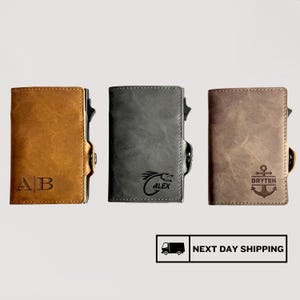 May include: Three leather wallets in shades of brown and grey. The first wallet is tan with the initials "AB". The second wallet is grey with a dragon graphic and the name "ALEX". The third wallet is brown with an anchor graphic and the name "DRYTEN".