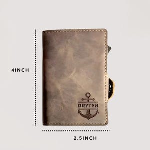 May include: A grey leather wallet, 10 cm tall and 6.35 cm wide, featuring an anchor design and the name "BRYTEN". The wallet has a stitched border and a card slot.