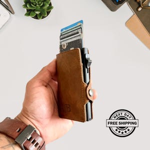 May include: A brown leather wallet with a black mechanism and a button closure. The wallet is holding several credit cards. The initials "AB" are embossed on the front. The watch band is brown leather.