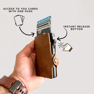 May include: A brown leather wallet with a black metal card holder. The wallet has the initials "AB" embossed on the front. The text reads "ACCESS TO YOU CARDS WITH ONE PUSH" and "INSTANT RELEASE BOTTON".