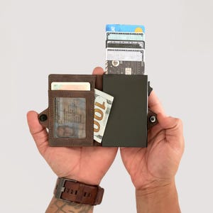 May include: A brown leather wallet with a dark grey metal card holder. The wallet is open, revealing a clear ID window, a &pound;80 note, and several credit cards. The wallet is held in two hands.
