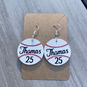May include: A pair of white baseball-themed earrings with red stitching. The earrings feature the name "Thomas" and the number "25" in black lettering.