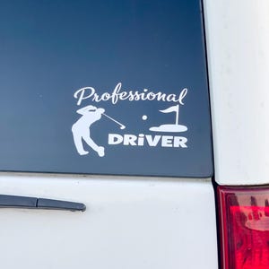 May include: White vinyl decal featuring a golfer in action, a golf ball, and a flag on a green. The words "Professional Driver" are written in a stylish font above and below the golfer illustration. The decal is affixed to a dark blue surface.