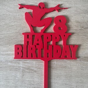 Puede incluir: Un adorno rojo para pastel con la silueta de un superh&eacute;roe, las palabras "HAPPY BIRTHAY" y el n&uacute;mero "8". Este adorno est&aacute; dise&ntilde;ado para una celebraci&oacute;n de cumplea&ntilde;os.