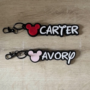 May include: Two keychains with black hardware and the names "CARTER" and "AVORY" in white, stylized lettering. Each keychain features a Mickey Mouse head silhouette in red and pink, respectively, on a black background.