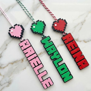 May include: Three personalized name tags with pixelated heart designs. One tag is pink and says "ARIELLE", another is green and says "HAYLON", and the last is red and says "REINA". Each tag is attached to a string.