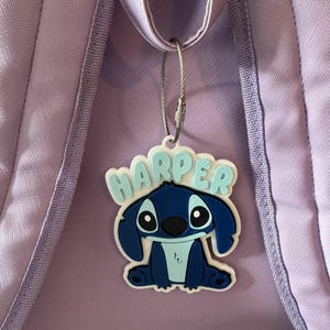 May include: A blue and white cartoon character luggage tag with the name "HARPER" in light blue letters. The tag is attached to a purple backpack with a silver wire loop.
