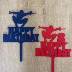 Puede incluir: Dos adornos para tartas con la silueta de un superh&eacute;roe. Uno es azul con el texto "HAPPY BIRTHAY", el otro es rojo con "HAPPY BIRTHDAY" y el n&uacute;mero "8". Ambos tienen un palito para insertar.