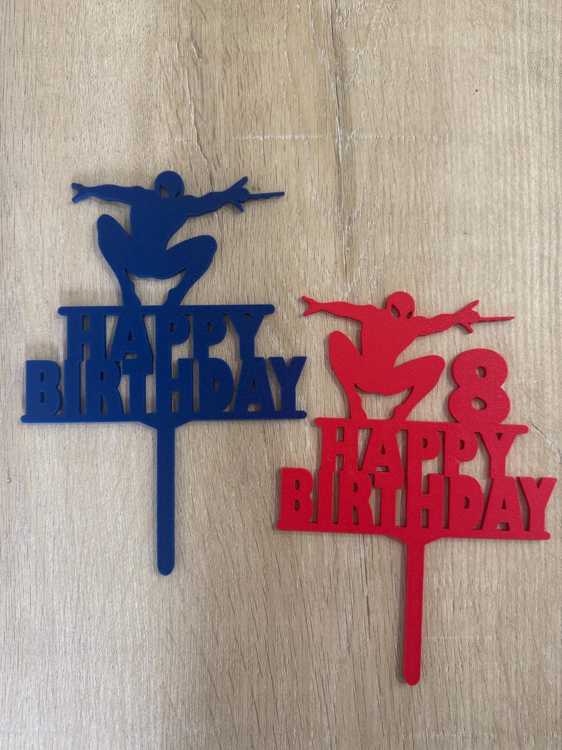 Puede incluir: Dos adornos para tartas con la silueta de un superh&eacute;roe. Uno es azul con el texto "HAPPY BIRTHAY", el otro es rojo con "HAPPY BIRTHDAY" y el n&uacute;mero "8". Ambos tienen un palito para insertar.