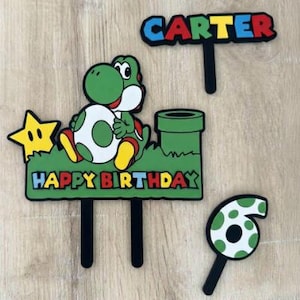May include: A set of colourful cake toppers with a Yoshi character, a star, a green pipe, and the words "HAPPY BIRTHDAY" and "CARTER". Also includes a number 6 topper. The toppers have black stems for easy cake insertion.