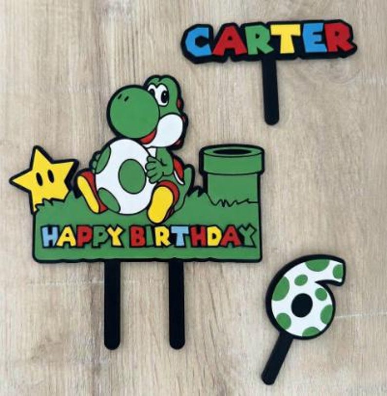 May include: A set of colourful cake toppers with a Yoshi character, a star, a green pipe, and the words "HAPPY BIRTHDAY" and "CARTER". Also includes a number 6 topper. The toppers have black stems for easy cake insertion.