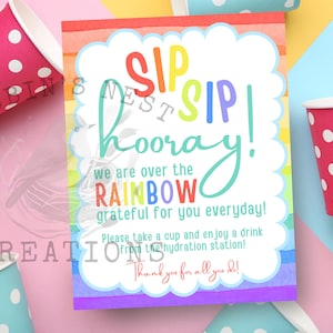 May include: A colorful sign with the words "SIP SIP hooray!" and "we are over the RAINBOW." Pink and blue polka dot cups surround the sign. The sign encourages people to take a cup and enjoy a drink from the hydration station.