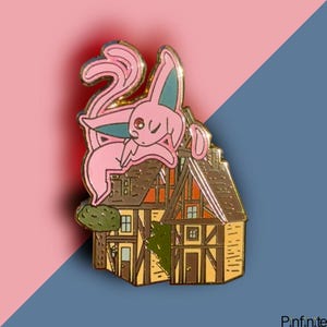 May include: Gold-tone enamel pin featuring a pink and blue Pokemon character, known as Espeon, perched on top of a small, brown, and yellow house with a green bush.