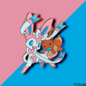 May include: A pink and blue enamel pin featuring a cartoon image of two Pokemon characters, Gardevoir and Eevee. Gardevoir is holding Eevee in her arms. The pin has a sparkly finish.