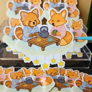 Puede incluir: Una colección de pegatinas con pandas rojos y zorros de dibujos animados con batas de colores pastel, tomando té en una plataforma con forma de nube. Cada pegatina tiene una pequeña mesa de madera con una tetera y tazas. Las pegatinas están decoradas con estrellas amarillas.