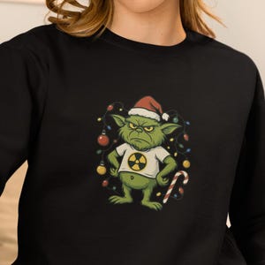 May include: Black sweatshirt featuring a green goblin wearing a Santa hat and a white t-shirt with a radiation symbol. The goblin is surrounded by Christmas lights and a candy cane. The sweatshirt is a crew neck style.