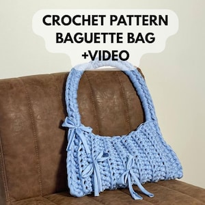 Puede incluir: Un bolso baguette de ganchillo azul claro con un asa redondeada y lazos decorativos. El bolso se muestra sobre una silla de cuero marrón. El texto en la parte superior dice "CROCHET PATTERN BAGUETTE BAG +VIDEO".
