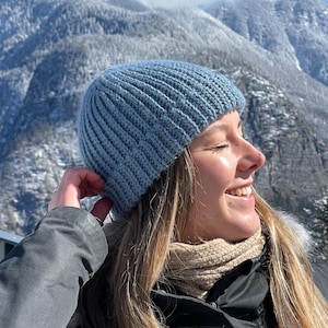 May include: A woman wearing a light blue knitted beanie, a beige scarf, and a black jacket, smiling and looking towards the snowy mountains in the background.