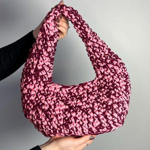 May include: A handmade, pink and burgundy crochet hobo bag. The bag is held up by two hands. The bag has a curved shape with a thick strap and a textured, chunky stitch pattern. The color combination is a mix of light pink and dark burgundy.