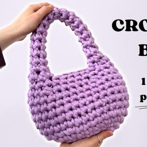 Op de afbeelding: Een lavendelkleurige gehaakte tas met één handvat. De tas is klein en zou perfect zijn om kleine spullen in te dragen. De tekst "CROCHET BAG 1 hour project" is zichtbaar in de afbeelding.